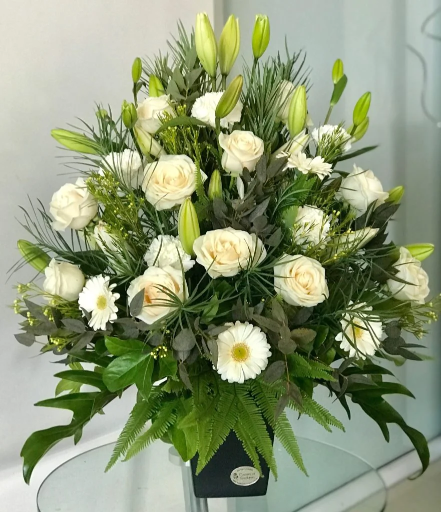 white flower arrangement in black vase with lilies roses and daisies on nude background