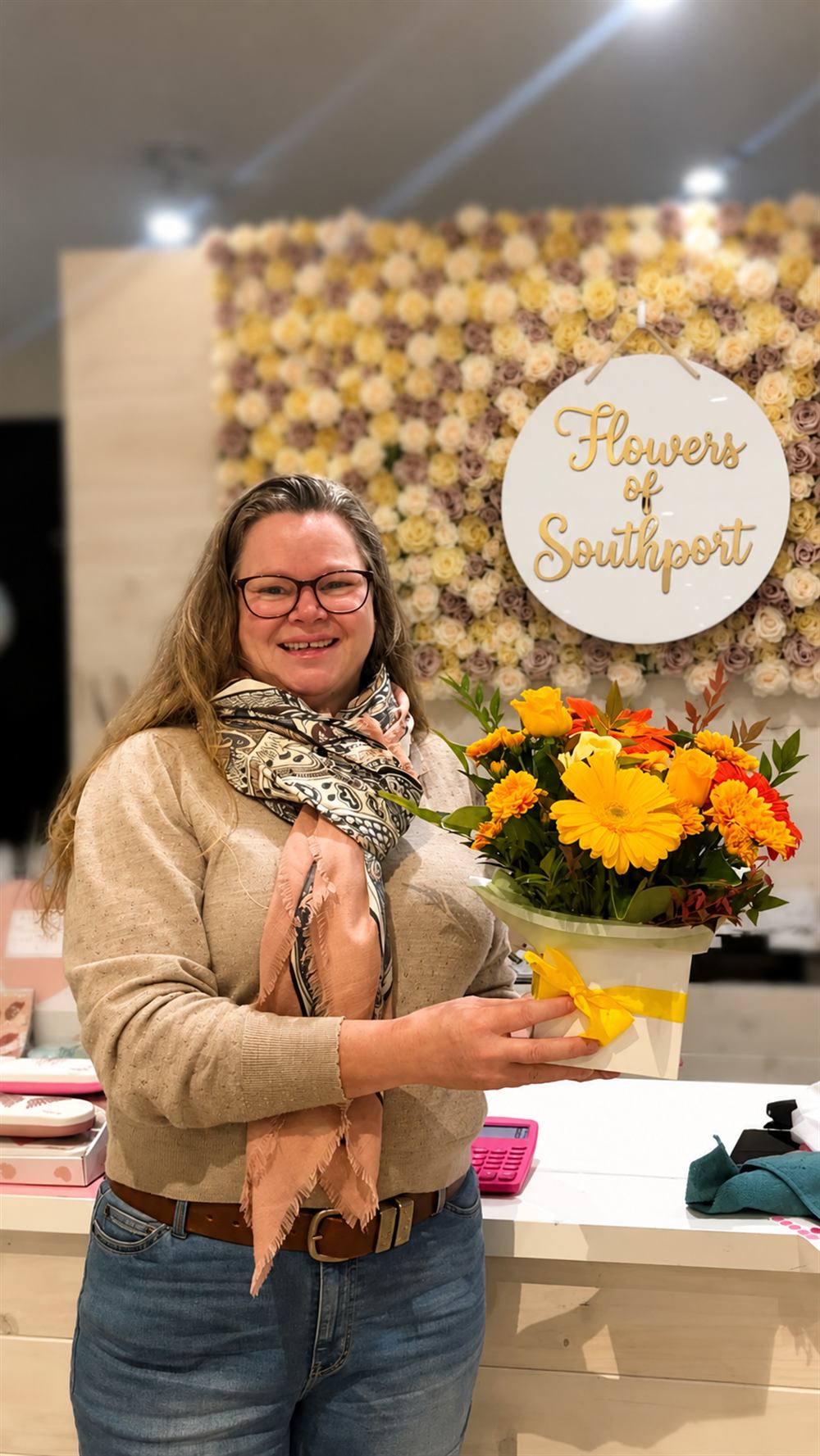 Ailsa, our second-place Mother's Day giveaway winner, holding her warm yellow Flowers of Southport arrangement in front of the rose wall at our Southport shop.