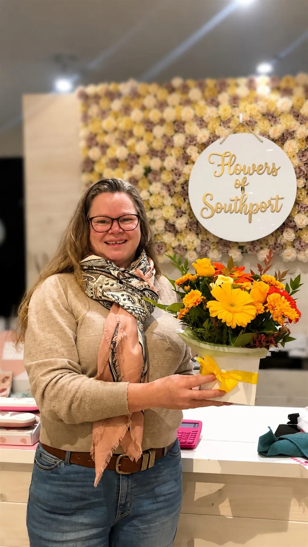 Ailsa, our second-place Mother's Day giveaway winner, holding her warm yellow Flowers of Southport arrangement in front of the rose wall at our Southport shop.