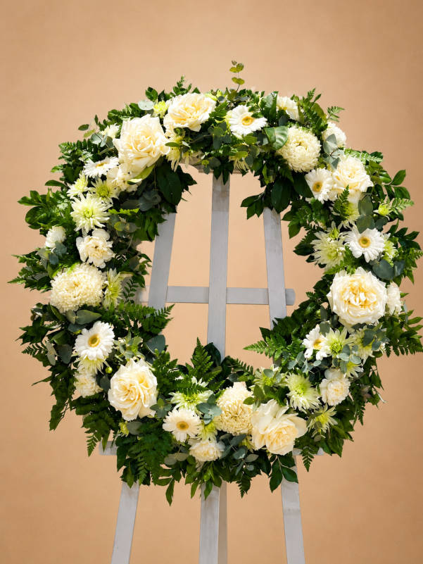 White funeral wreath of roses, gerberas and chrysanthemums with eucalyptus and fern foliage, displayed on an easel.